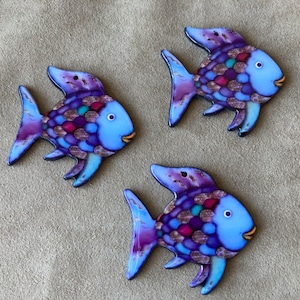 May include: Three blue ceramic fish ornaments with purple, pink, and turquoise scales. The fish have a whimsical, cartoon-like design.