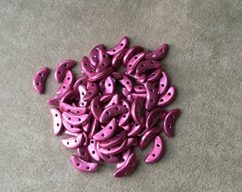 May include: A pile of metallic pink crescent-shaped beads with two holes each. The beads are scattered on a neutral-toned surface, creating a textured appearance. These beads are ideal for jewelry making and craft projects.