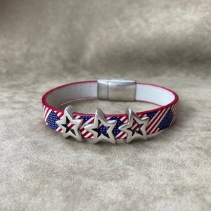 May include: A patriotic bracelet featuring the American flag design with red and white stripes and blue star accents. The bracelet has a white interior and silver-colored star charms. The bracelet is approximately 0.75 inches wide.