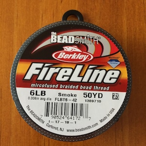 May include: A spool of black FireLine microfused braided bead thread by Berkley. The spool is labeled with the text "6LB Smoke 50YD" and a barcode. The text "The Beadsmith Carteret, NJ www.beadsmith.com Made in USA" is printed on the spool.