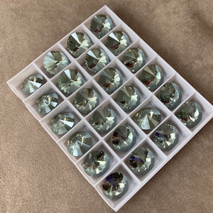 May include: A white plastic tray with 25 clear glass rhinestones. The rhinestones are arranged in a grid pattern and have a silver-grey color.
