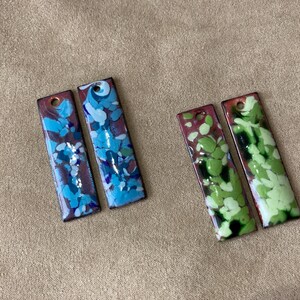 May include: Four rectangular pendants with a hole at the top. Two pendants feature blue and purple abstract designs, while the other two display green and white patterns. The pendants have a dark border and are set against a neutral background.