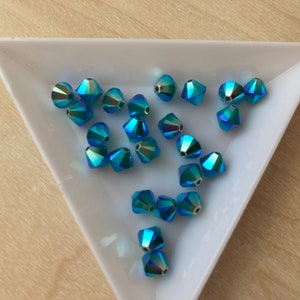 May include: A collection of teal blue iridescent glass beads in a triangular white dish. The beads are faceted and have a pointed shape.