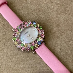 May include: A pink leather strap watch with a silver bezel and a mother-of-pearl face. The watch face is surrounded by colorful gemstones. The watch is branded "PEDRE".