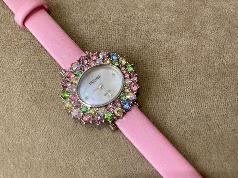 May include: A pink leather strap watch with a silver bezel and a mother-of-pearl face. The watch face is surrounded by colorful gemstones. The watch is branded "PEDRE".