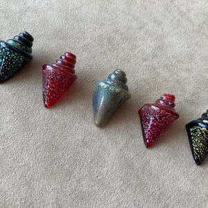May include: Five colorful glass cone-shaped beads with spiral tops. The beads are in shades of red, green, and black, with speckled and iridescent finishes. The beads are arranged diagonally on a neutral surface.