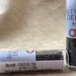 May include: Two small clear glass bottles filled with black seed beads. The bottles have white labels with black text. The label on the left bottle reads "Item#: DB310-TB APRX 7.2 GM DELICA BEAD MATTE BLACK". The label on the right bottle reads "MIYUKI DELICA 11/0" with a red circle and square.