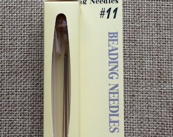 May include: A pack of four Tulip Beading Needles size #11 in a clear glass vial. The needles are gold in color and are packaged in a yellow cardboard box with a red tulip logo.