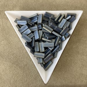 May include: A collection of metallic blue rectangular beads with a slight iridescent sheen, filling a white triangular dish. The beads are of varying sizes and shapes, suggesting a handmade or artisanal quality. The background is a neutral tan fabric.