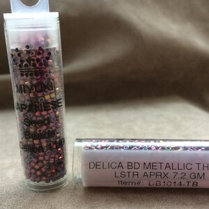 May include: Two small clear plastic tubes filled with metallic seed beads. The tube on the left has a label that reads "MIYUKI JAPANESE Seed Beads Delica 11/0". The tube on the right has a label that reads "DELICA BD METALLIC THISTLE LSTR APRX 7.2 GM Item#: DB1014-TB".