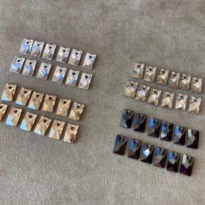 May include: An assortment of rectangular crystal rhinestones in various colors, including clear, gold, and dark gray. Each rhinestone has a flat back and a hole at the top for attaching to fabric or other surfaces. The crystals are arranged in neat rows.
