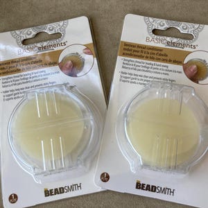 May include: Two packages of Basic Elements beeswax thread conditioner by The Beadsmith. Each package contains a round, pale yellow disc of beeswax housed in a clear plastic holder with a hinged lid, designed for beading and hand sewing.