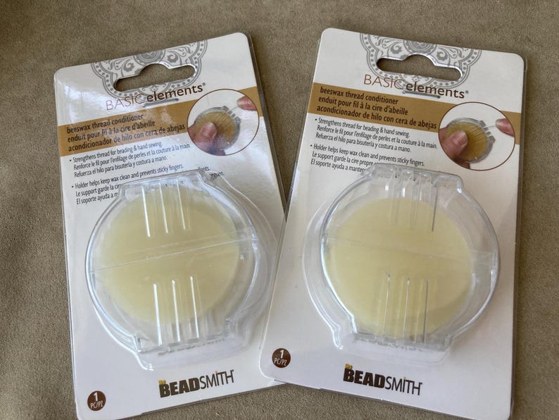 May include: Two packages of Basic Elements beeswax thread conditioner by The Beadsmith. Each package contains a round, pale yellow disc of beeswax housed in a clear plastic holder with a hinged lid, designed for beading and hand sewing.