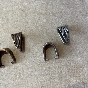 May include: Four metal jewelry bails arranged in a group. Two bails have a bronze-toned finish and two bails have a silver-toned finish. Each bail features a curved, leaf-like embossed pattern on the exterior surface and two small prongs.