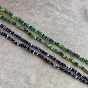 May include: Three strands of square glass beads. The top strand is green with white and black swirls. The middle strand is green with white swirls. The bottom strand is black with white swirls.