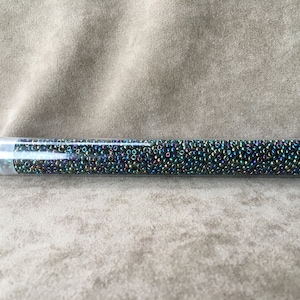 Op de afbeelding: Een transparante plastic buis gevuld met kleine, iriserende zwarte, groene en blauwe zaadkraaltjes. De buis heeft een wit label met het nummer "11-9455" erop.