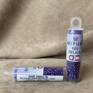 May include: Two small clear plastic tubes filled with purple seed beads. The tubes have labels that read "MIYUKI 11/0 DELICA" and "Item#: DB923-TB DELICA BD SPRKLNG VIOLET LINED CRYSTAL- APRX 7.2GM".