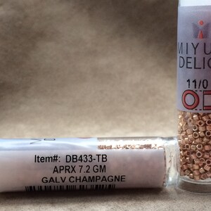 May include: Two small glass bottles filled with gold-colored seed beads. The bottle on the left has a white label with the text "Item#: DB433-TB APRX 7.2 GM GALV CHAMPAGNE". The bottle on the right has a white label with the text "MIYUKI DELICA 11/0".