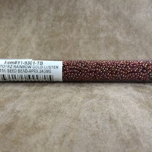 May include: A clear plastic tube filled with small, iridescent, dark topaz rainbow gold luster seed beads. The tube has a label that reads "Item#11-9301-TB DK TOPAZ RAINBOW GOLD LUSTER 11/0 SEED BEAD-APRX 24GMS".