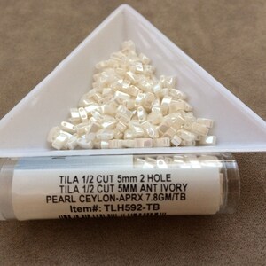 May include: A small container of white, 5mm, two-hole, half-cut glass beads. The label reads "TILA 1/2 CUT 5mm 2 HOLE TILA 1/2 CUT 5MM ANT IVORY PEARL CEYLON-APRX 7.8GM/TB Item#: TLH592-TB".