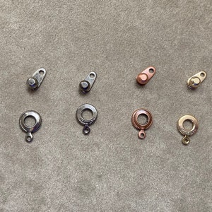 May include: An assortment of jewelry clasps in various metallic finishes, including silver, gunmetal, copper, and gold. The clasps are in different shapes and sizes, with some featuring a circular design and others a more rectangular shape.