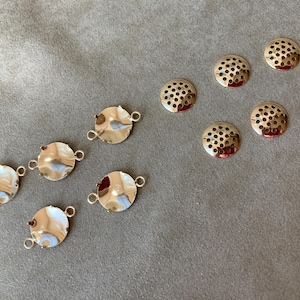 May include: An assortment of jewelry findings, including several silver-toned, round connector pieces with small loops and a set of round, perforated beads. The findings are arranged on a gray surface.