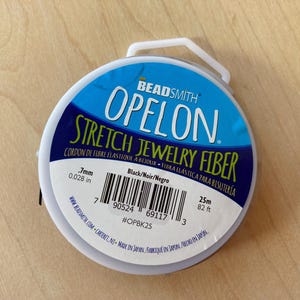 May include: A spool of black elastic stretch jewelry fiber, labeled "Beadsmith Opelon Stretch Jewelry Fiber" with the text "Cordon de fibre elastique a bijoux - Fibra elastica para bisuteria" in French and Spanish. The spool is 25 meters (82 feet) long and 0.7 mm (0.028 inches) thick. The label also includes a barcode and the product number #OPBK25.