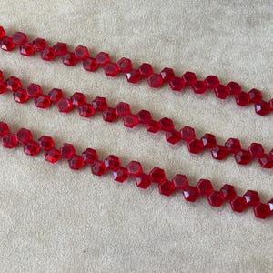 May include: Three strands of red, faceted beads arranged diagonally across a neutral background. The beads are hexagonal in shape and strung together, creating a zig-zag pattern. The beads are a deep, transparent red.