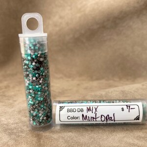 May include: Two small clear plastic tubes filled with seed beads. The beads are a mix of silver, black, and turquoise. The tube on the right has a label that reads "BBD DB MIX Color: Mint Opal $7-".