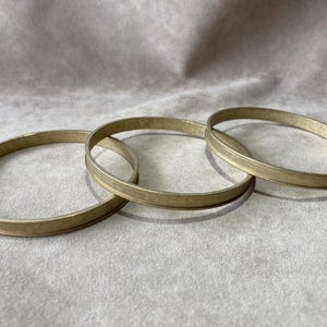 May include: Three gold-colored bangles are arranged on a light gray surface. The bangles are circular, with a flat, slightly wider band. The metal has a matte finish, and the bangles are stacked slightly, creating shadows.