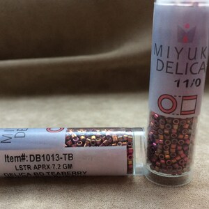 Könnte beinhalten: Zwei kleine Glasbehälter gefüllt mit kupfernen und goldenen Perlen. Die Behälter sind mit "MIYUKI DELICA 11/0" und "Item#: DB1013-TB LSTR APRX 7.2 GM DELICA BD TEABERRY" beschriftet.