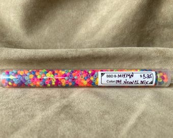 Puede incluir: Un tubo de plástico transparente lleno de cuentas de semillas de colores. La etiqueta dice "BBD 8 - MIXMN 5,45 € Color: Mt. Neon CL Mix".