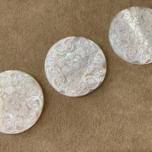 May include: Three round, white, mother-of-pearl buttons with intricate floral and scrollwork designs. The buttons are arranged on a brown, textured surface, showcasing their pearlescent sheen and detailed carvings.