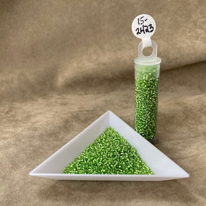 May include: A small container of green seed beads, labeled "15-2423", is displayed on a white triangular tray. The beads are arranged in a pile on the tray.