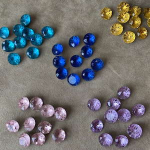 Op de afbeelding: Assortiment van gefacetteerde glazen edelstenen in verschillende kleuren, waaronder blauw, paars en goud. De edelstenen zijn in groepen gerangschikt op een getextureerd grijs oppervlak.