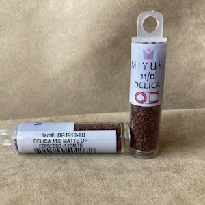May include: Two clear plastic tubes of Miyuki Delica beads. One tube is upright, the other is lying down. The tubes contain small, matte espresso-colored beads. The labels read "MIYUKI 11/0 DELICA" and "DELICA 11/0 MATTE OP ESPRESSO".