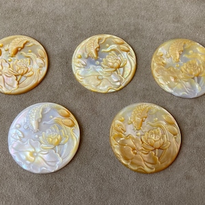 May include: Five round, iridescent mother-of-pearl cabochons. Each features a carved design of a koi fish swimming above a lotus flower. The colors range from cream to gold, with subtle variations in the pearl's sheen. The cabochons are set against a neutral background.