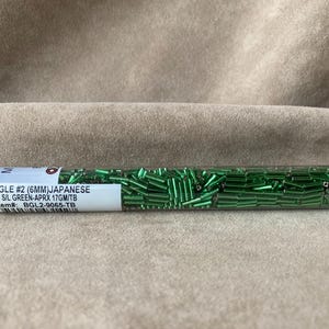 May include: A close-up of a clear plastic tube filled with green glass bugle beads. The label reads "BUGLE #2 (6MM)JAPANESE SIL GREEN-APRX 17GM/TB Item#: BGL2-9065-TB".