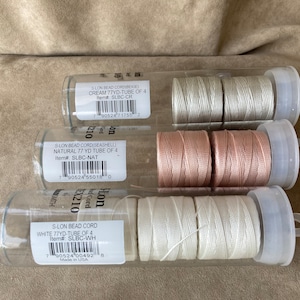 以下が含まれることがあります： ビーズコードが巻かれた透明なプラスチック製のチューブが3本。チューブにはコードの色と長さが表示されています。色はクリーム、ナチュラル、ホワイトです。各チューブにはコードが4巻き入っています。