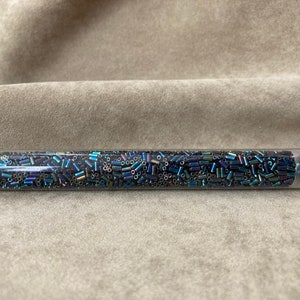 Op de afbeelding: Een transparante plastic buis gevuld met iriserende blauwe en zwarte kralen. De buis heeft een wit label met de tekst "BGH-9455".