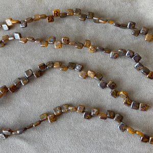 May include: Four strands of brown and gold square beads with intricate patterns. The beads are strung on clear thread.