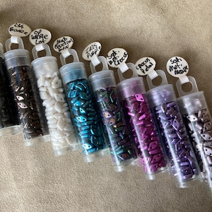 Może przedstawiać: Dziesięć małych, przezroczystyх probówek wypełnionych różnokolorowymi szklanymi koralikami. Probówki są oznaczone kolorem i wykończeniem koralików. Kolory obejmują czarny, brązowy, biały, turkusowy, zielony, fioletowy i liliowy.