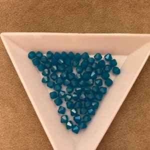 May include: A close-up of a white triangular tray filled with teal blue glass beads. The beads are faceted and have a shiny finish.