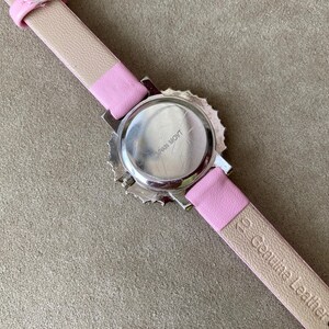 May include: A wristwatch with a pink leather strap and a silver-colored watch face. The back of the watch is visible, showing the words "Japan Movt". The strap is a light pink color with the words "Genuine Leather" printed on it.