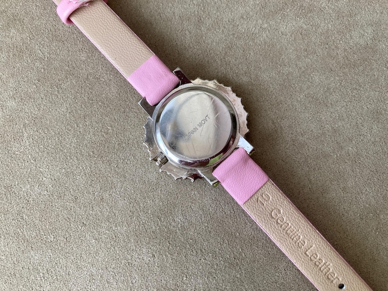 May include: A wristwatch with a pink leather strap and a silver-colored watch face. The back of the watch is visible, showing the words "Japan Movt". The strap is a light pink color with the words "Genuine Leather" printed on it.