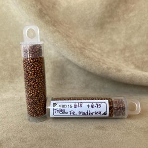 May include: Two clear plastic tubes filled with small, round, copper-colored beads. One tube stands upright, while the other lies horizontally. A label on the horizontal tube reads "Toho Color Fr. Mudbrick".