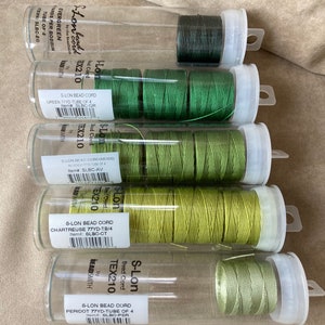 以下が含まれることがあります： エバーグリーン、チャートリュース、ペリドットなど、さまざまな緑色のシェードのS-Lonビーズコードが5個あります。各スプールには、色の名前、ヤード数、品番がラベル付けされています。スプールは、透明なプラスチック容器に垂直に配置されています。