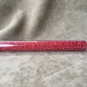 May include: A clear plastic tube filled with small, vibrant red beads. The beads have a slight iridescent sheen. The tube is sealed with a pink cap and contains a label with handwritten text. The beads are likely used for jewelry making or crafts.
