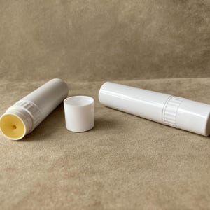 May include: Two white cylindrical lip balm tubes with a smooth plastic finish. One tube is closed with a cap, while the other is open, revealing a pale yellow balm core with a small central indentation. The tubes feature a ribbed twist mechanism.