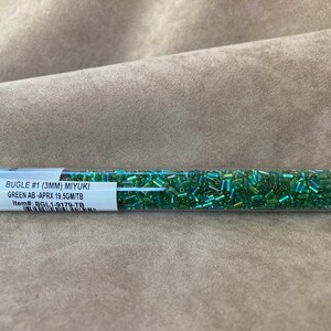 Op de afbeelding: Een doorzichtige plastic buis gevuld met groene bugle kralen met ab-afwerking. Het etiket vermeldt "BUGLE #1 (3MM) MIYUKI GREEN AB-APRX 19.5GM/TB Item# BGL1-9179-TB".